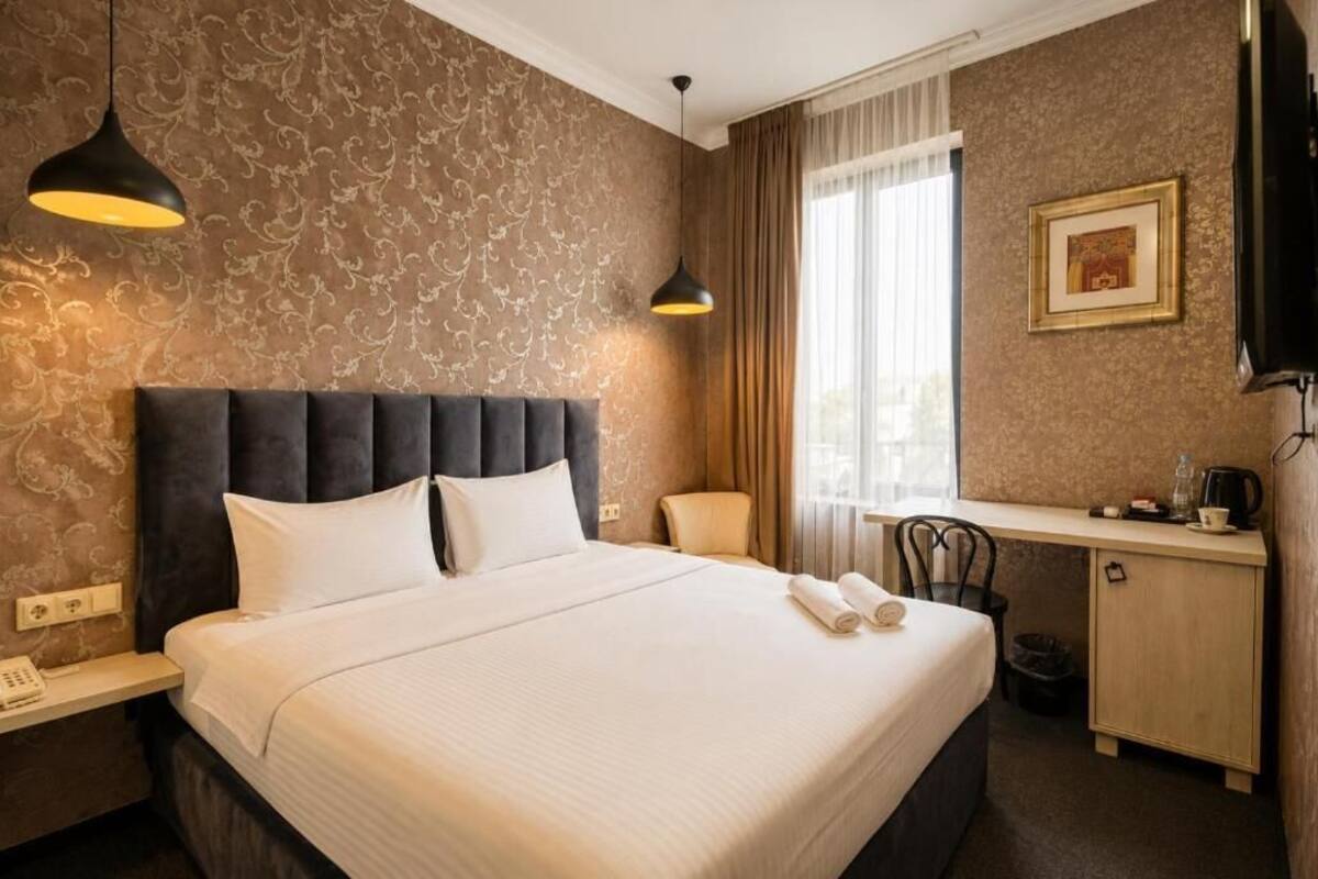 Hotellihuone, jossa on suuri sänky, kaksi riippuvalaisinta, työpöytä tuolilla, televisio ja seinällä oleva kehystetty taulu.