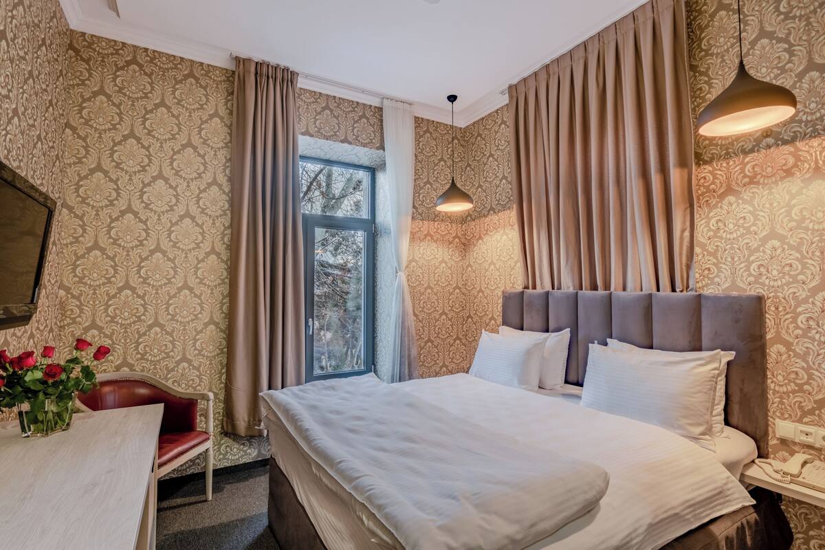 A hotel room with a bed, a desk, a chair, a vase of red roses, and a window with curtains.