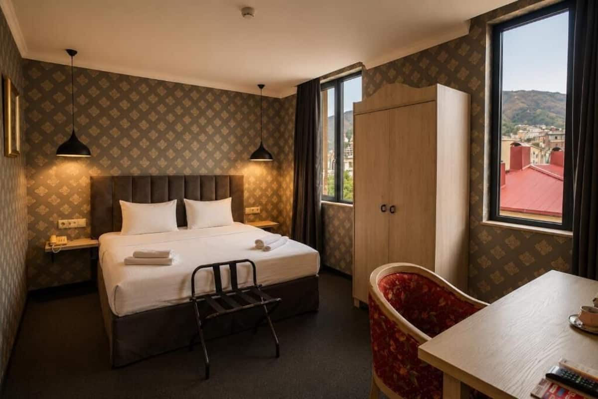 Hotellihuone, jossa on sänky, yöpöydät, työpöytä, tuoli, vaatekaappi ja ikkuna, josta on näkymä.