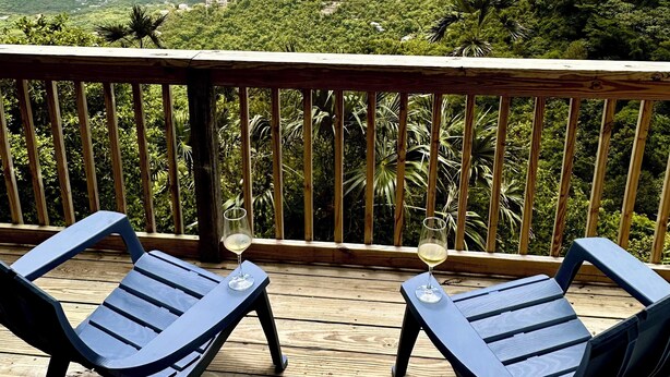 A wooden deck with two blue lounge chairs and a glass of white wine, overlooking a lush landscape with mountains and a turquoise bay. - NEW LISTING! Your Private Paradise on St. John’s Beautiful Bordeaux Mountain (St John)