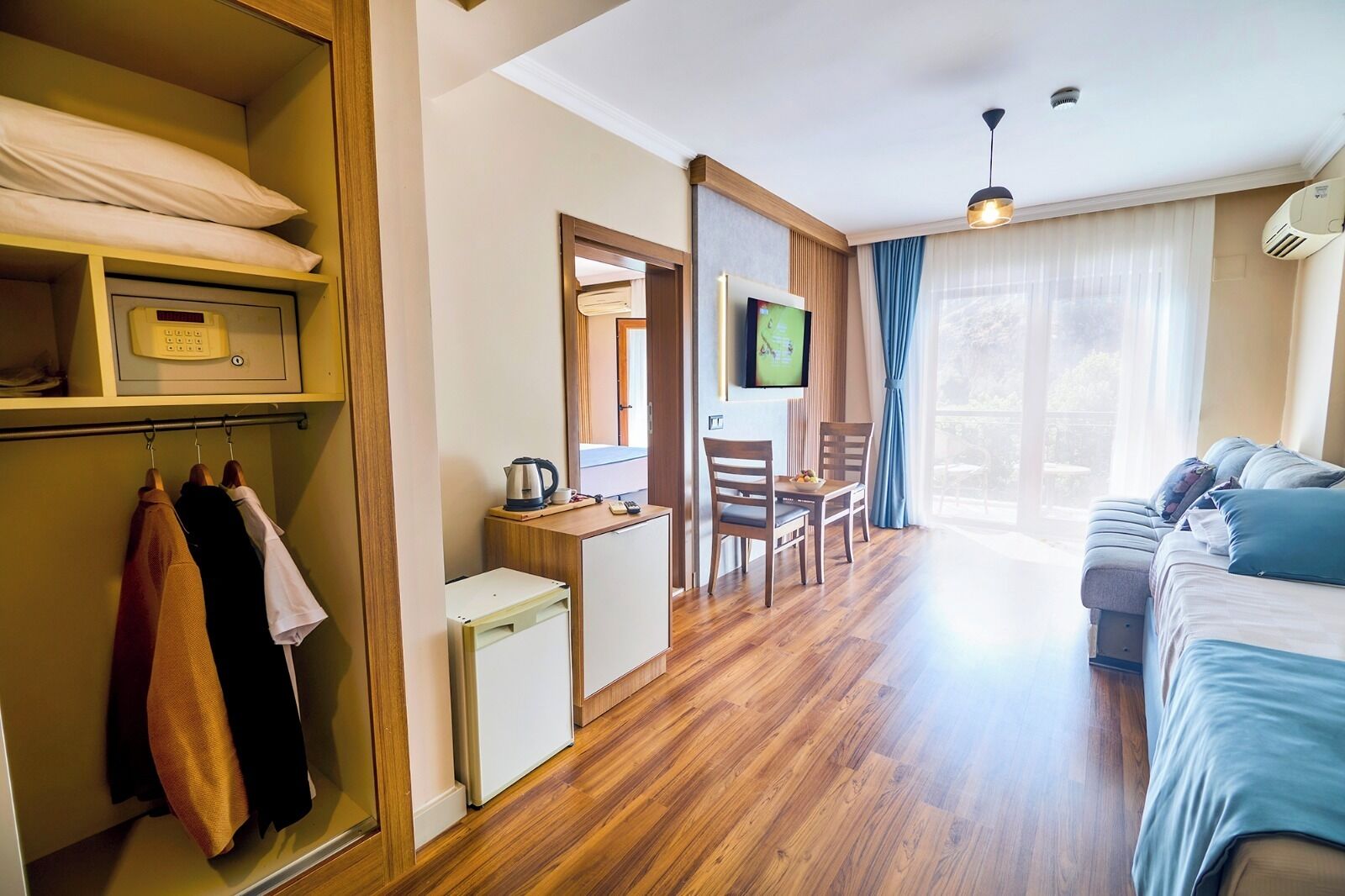 Ein Hotelzimmer mit Holzboden, einem Bett mit blauer Bettwäsche, einem an der Wand befestigten Fernseher, einem kleinen Tisch mit Wasserkocher und einem Kleiderschrank mit Kleidung.
