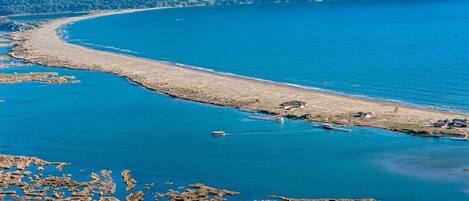An aerial view of a coastal area with a sandy beach, boats, and a forested mountain range in the background.