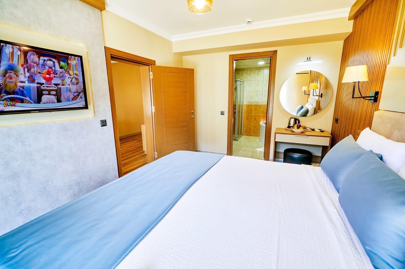 A hotel room with a bed, a desk with a mirror, a TV on the wall, and a bathroom visible through an open door.