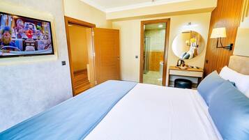 Habitación de hotel con cama, escritorio con espejo, televisor en la pared y baño a la vista a través de una puerta abierta.