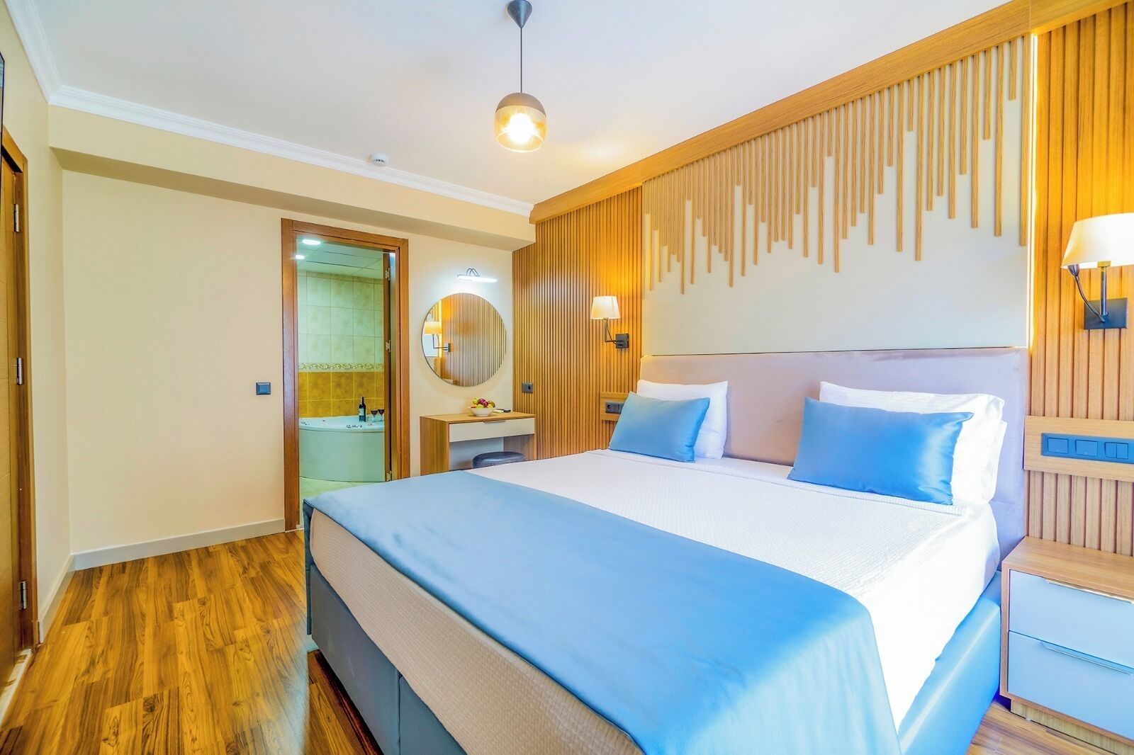 A hotel room with a large bed, wooden paneling on the walls, a round mirror, a bedside table with a lamp, and a bathroom visible through an open door.
