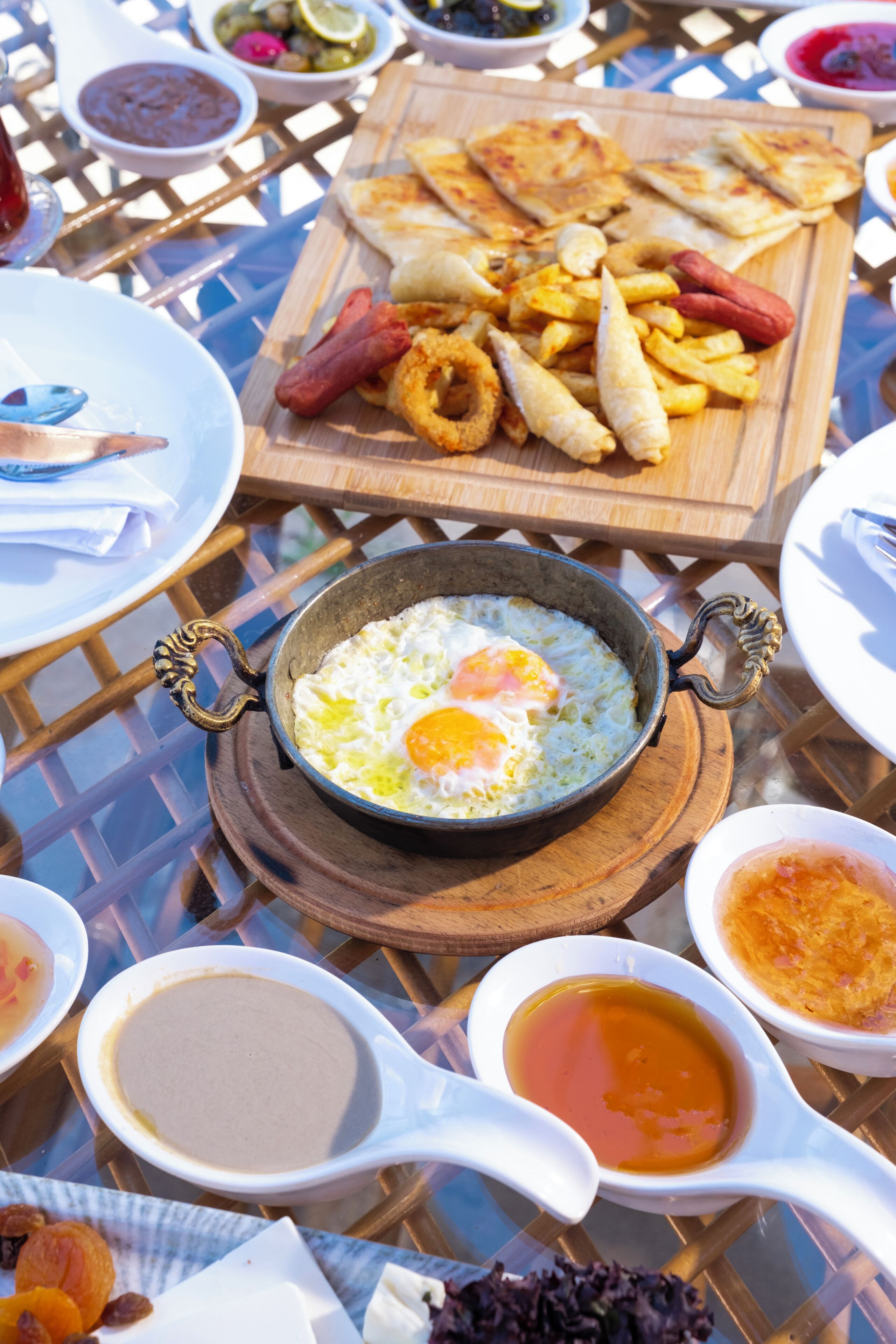 Una mesa de comedor con diversos platos, entre ellos huevos fritos, salchichas, y una tabla de madera con papas fritas y aros de cebolla.