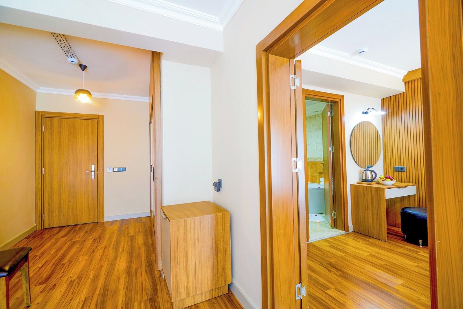 Une chambre d’hôtel avec un plancher en bois, un petit banc en bois, un luminaire au plafond, une armoire en bois et un miroir avec un cadre rond.
