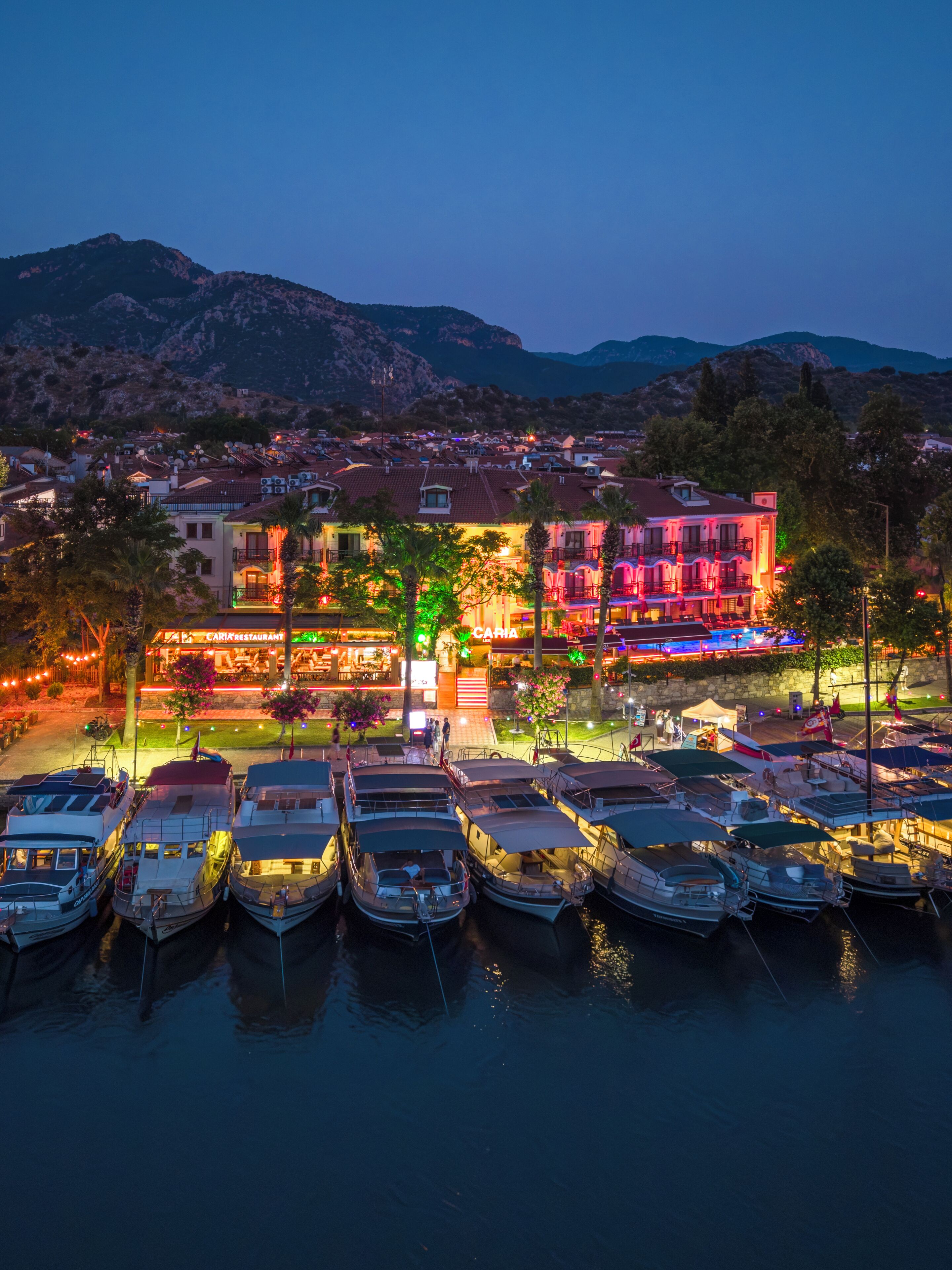 A marina with boats docked, a restaurant with outdoor seating, and a mountainous backdrop.