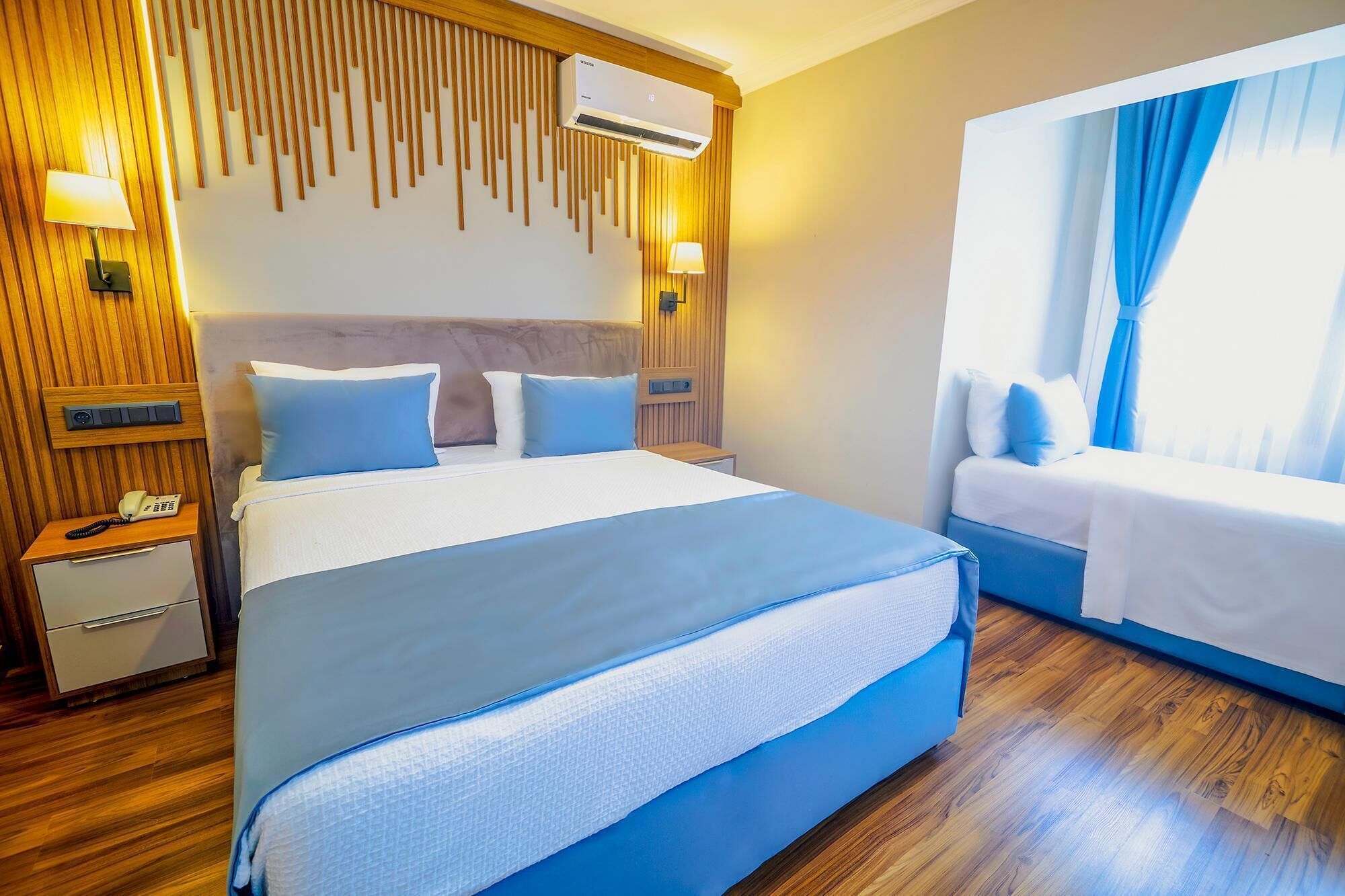 A hotel room with a large bed, a smaller bed, wooden paneling, a wall-mounted air conditioner, a bedside table with a phone, and a window with curtains.