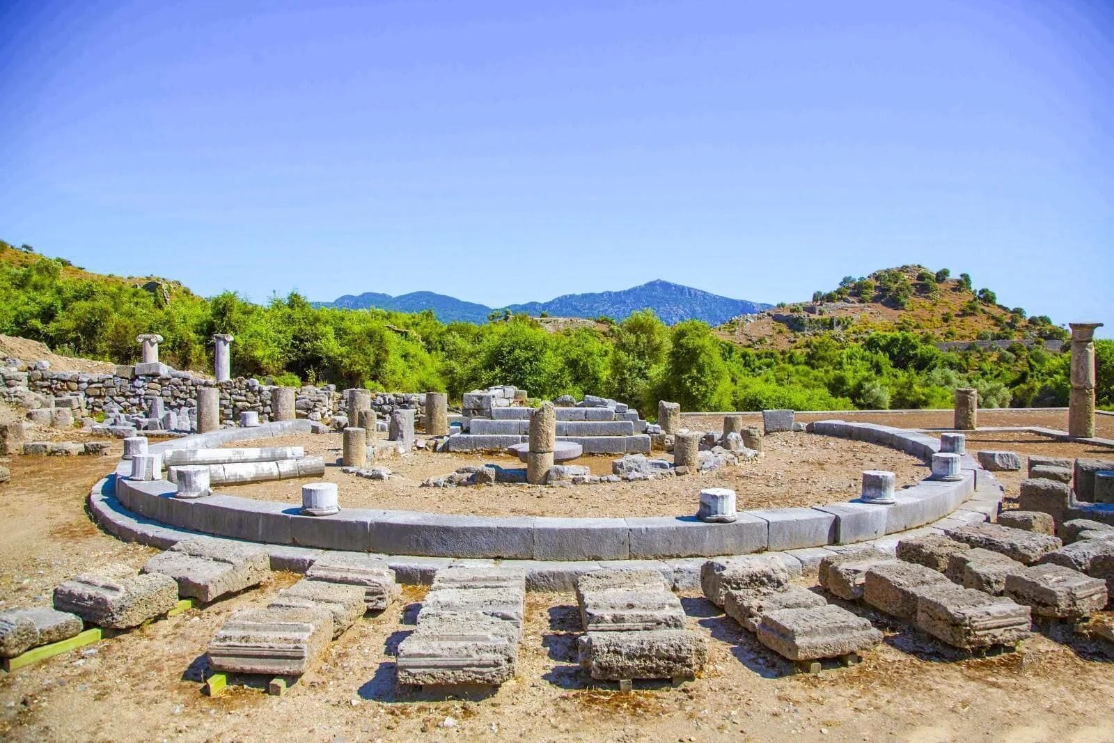 Ruinas antiguas con columnas de piedra y una estructura circular, rodeadas de vegetación y montañas al fondo.