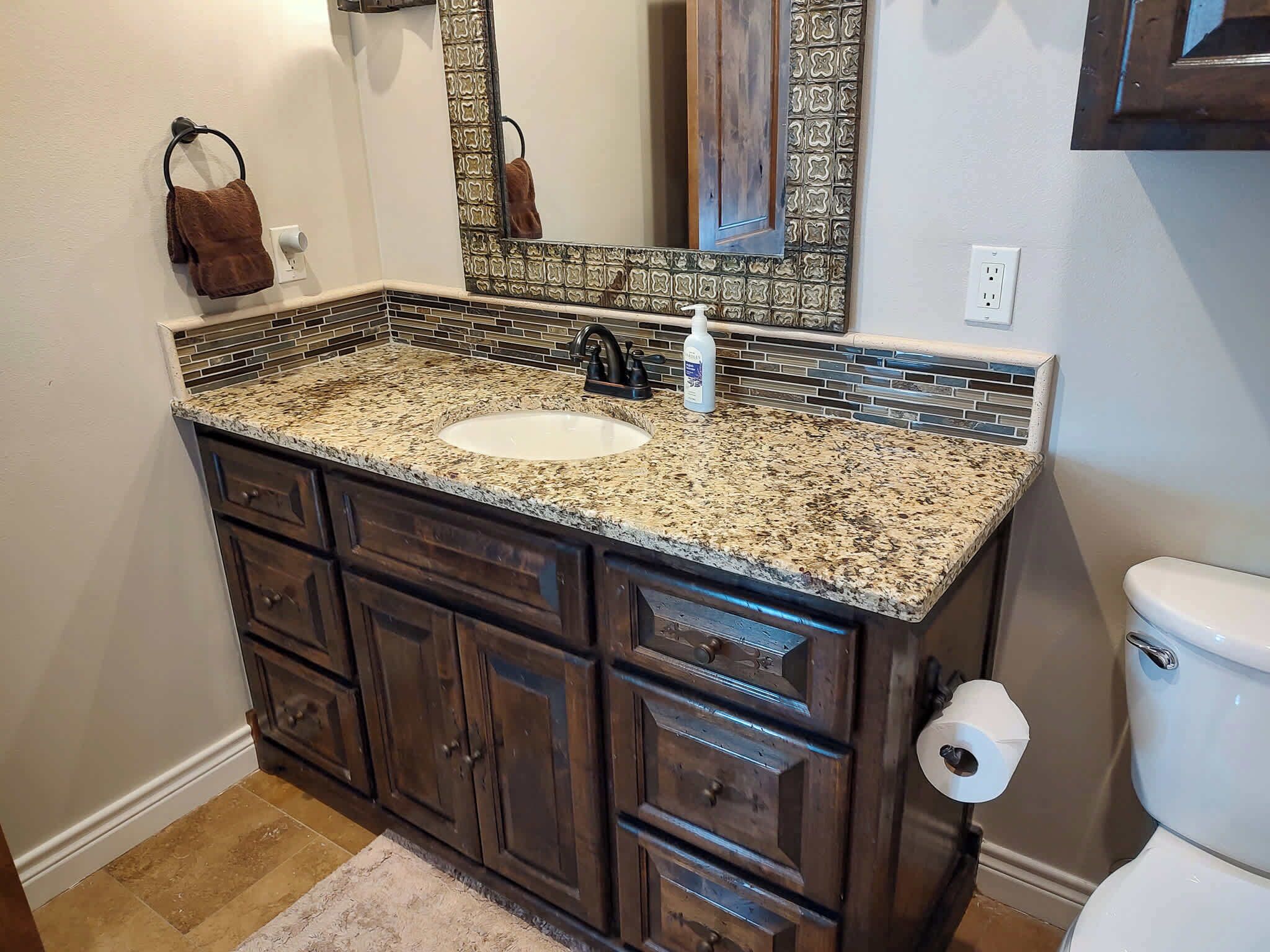 A bathroom with a granite countertop sink, dark wooden cabinets, a mirror, and a toilet.