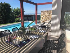 A poolside dining area with a table set for two, surrounded by chairs, a stone wall, and a view of a swimming pool and greenery.