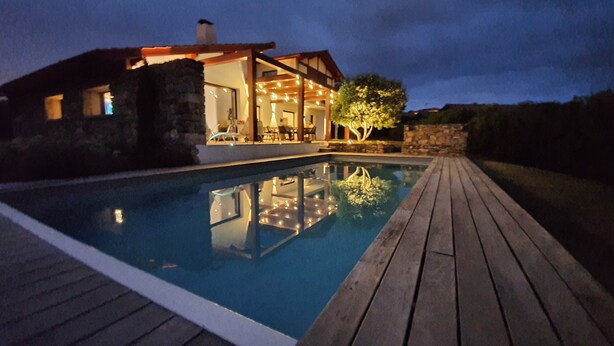 A swimming pool with a wooden deck, a stone wall, and a building with outdoor seating and string lights.