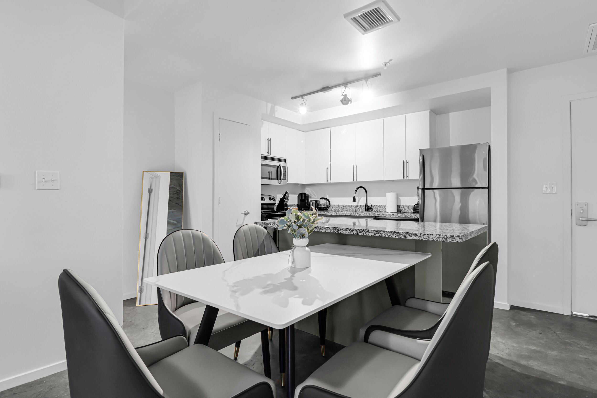 A modern kitchen with white cabinets, a stainless steel refrigerator, and a dining area with a white table and grey chairs.