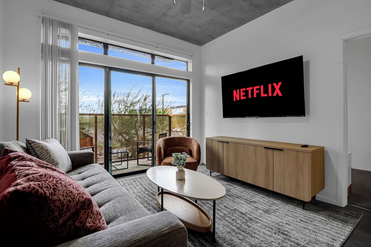 Ein modernes Wohnzimmer mit großem Fernseher, der "NETFLIX" zeigt, einer Couch, einem Couchtisch und einer Glasschiebetür, die auf einen Balkon führt.