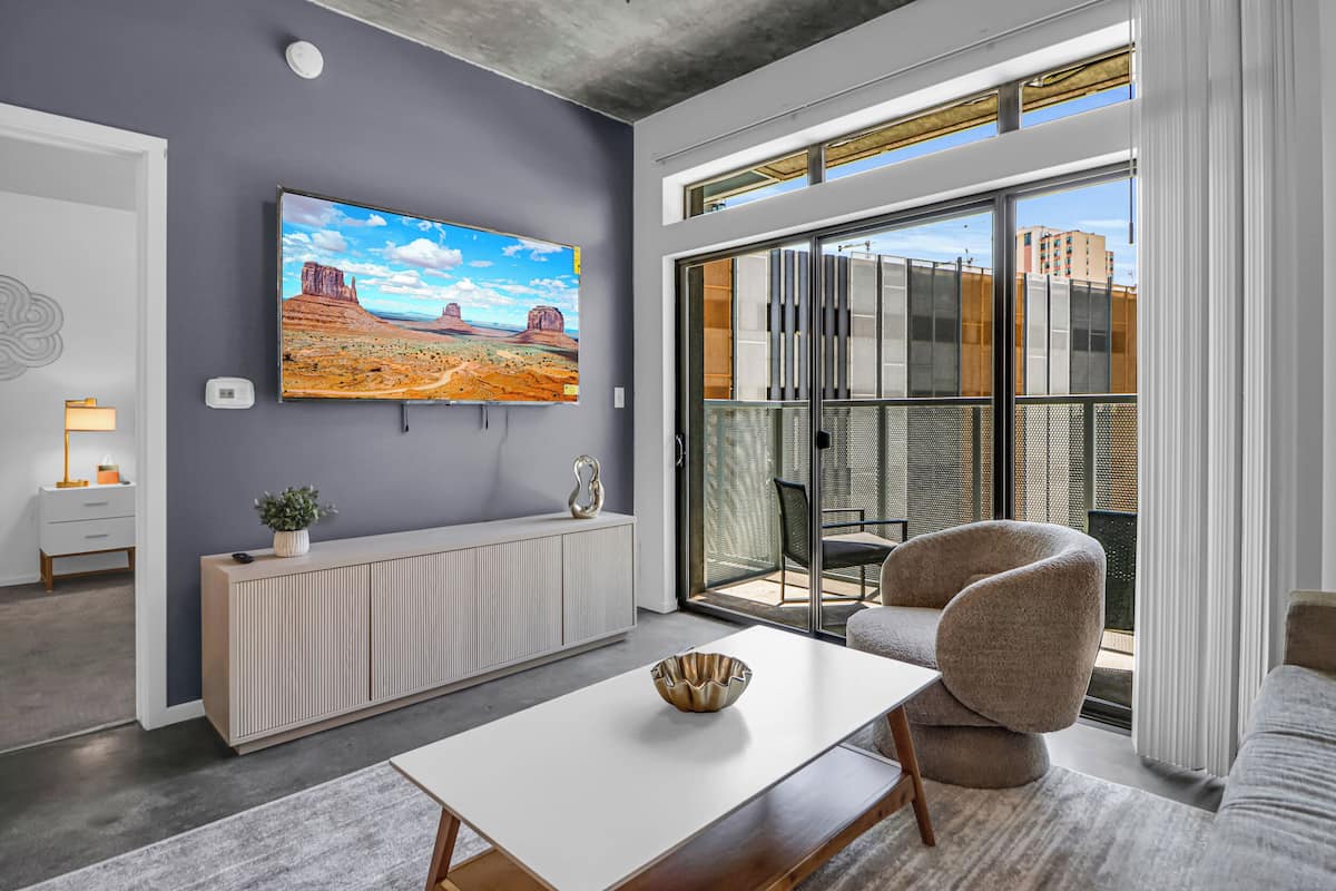 Ein modernes Wohnzimmer mit großem Fernseher, einer Couch, einem Esstisch und Blick auf eine Stadtkulisse durch große Fenster.