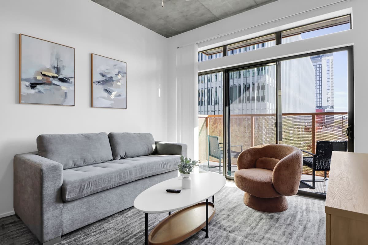 Ein modernes Wohnzimmer mit einem grauen Sofa, einem runden Couchtisch und einem braunen Sessel. An der Wand befinden sich zwei abstrakte Gemälde und ein großes Fenster mit Blick auf Gebäude.