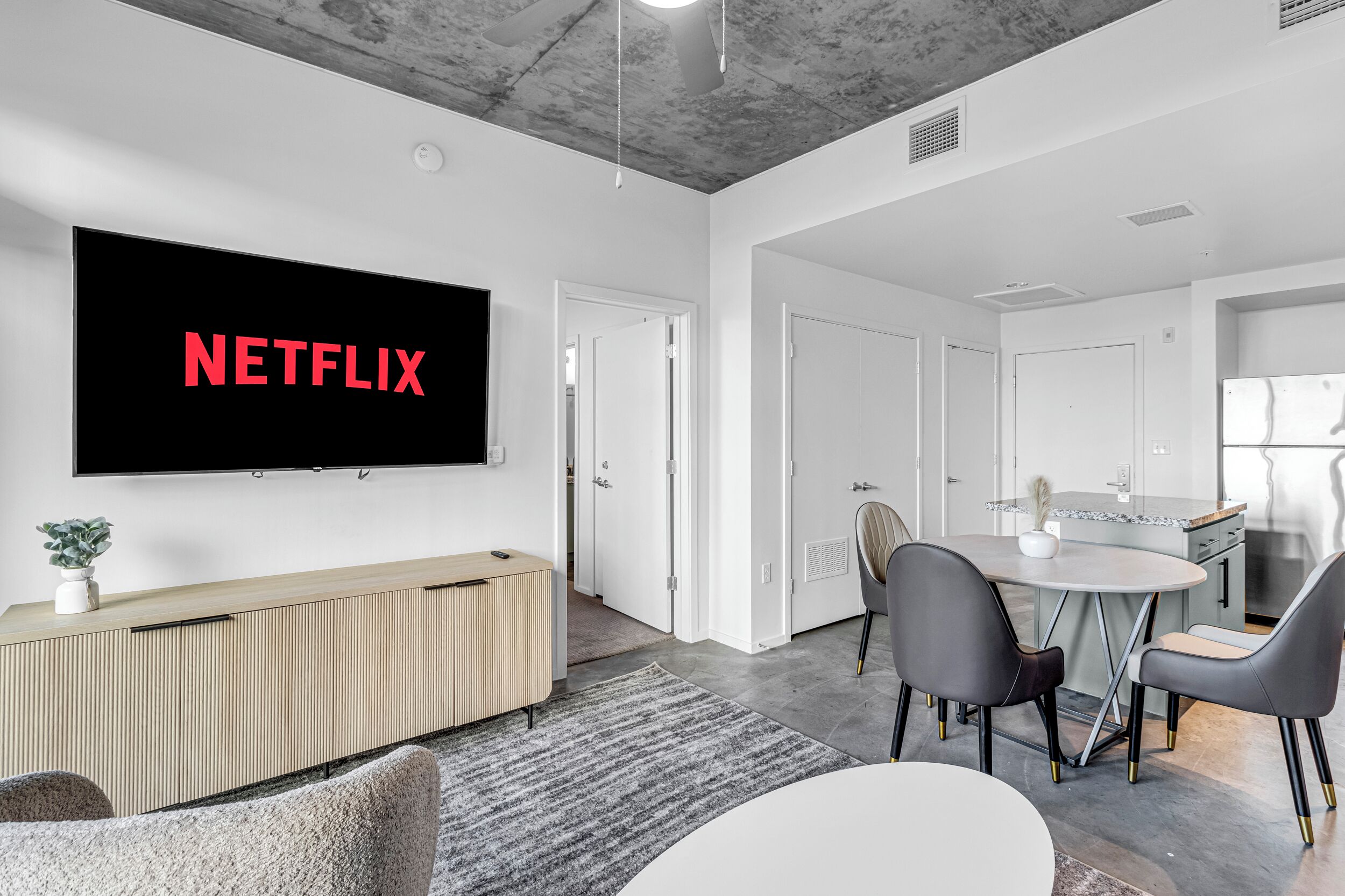 A modern living room with a flat-screen TV displaying "NETFLIX," a wooden console table, a gray sofa, a dining table with chairs, and a kitchen area.