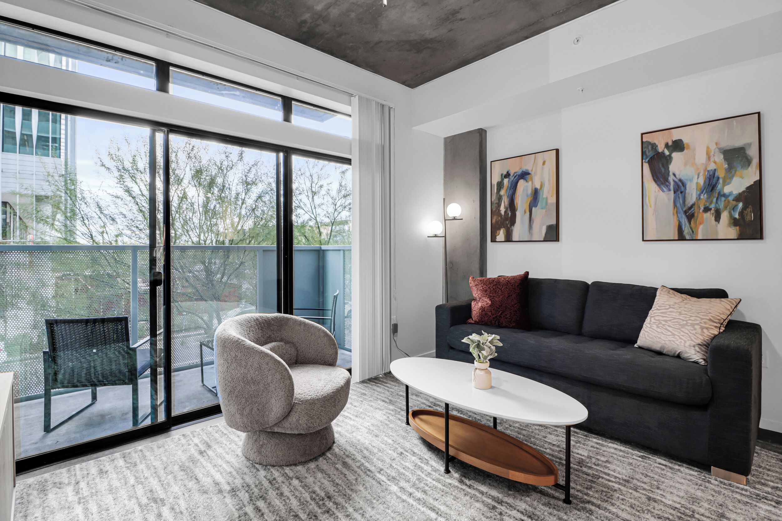 Un salon moderne avec un canapé, un fauteuil et une table basse, de grandes fenêtres et une œuvre d’art abstraite au mur.
