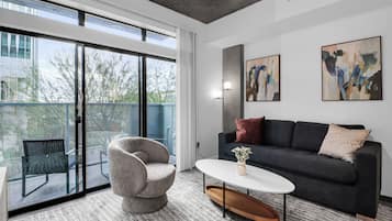 Un salon moderne avec un canapé, un fauteuil et une table basse, de grandes fenêtres et une œuvre d’art abstraite au mur.