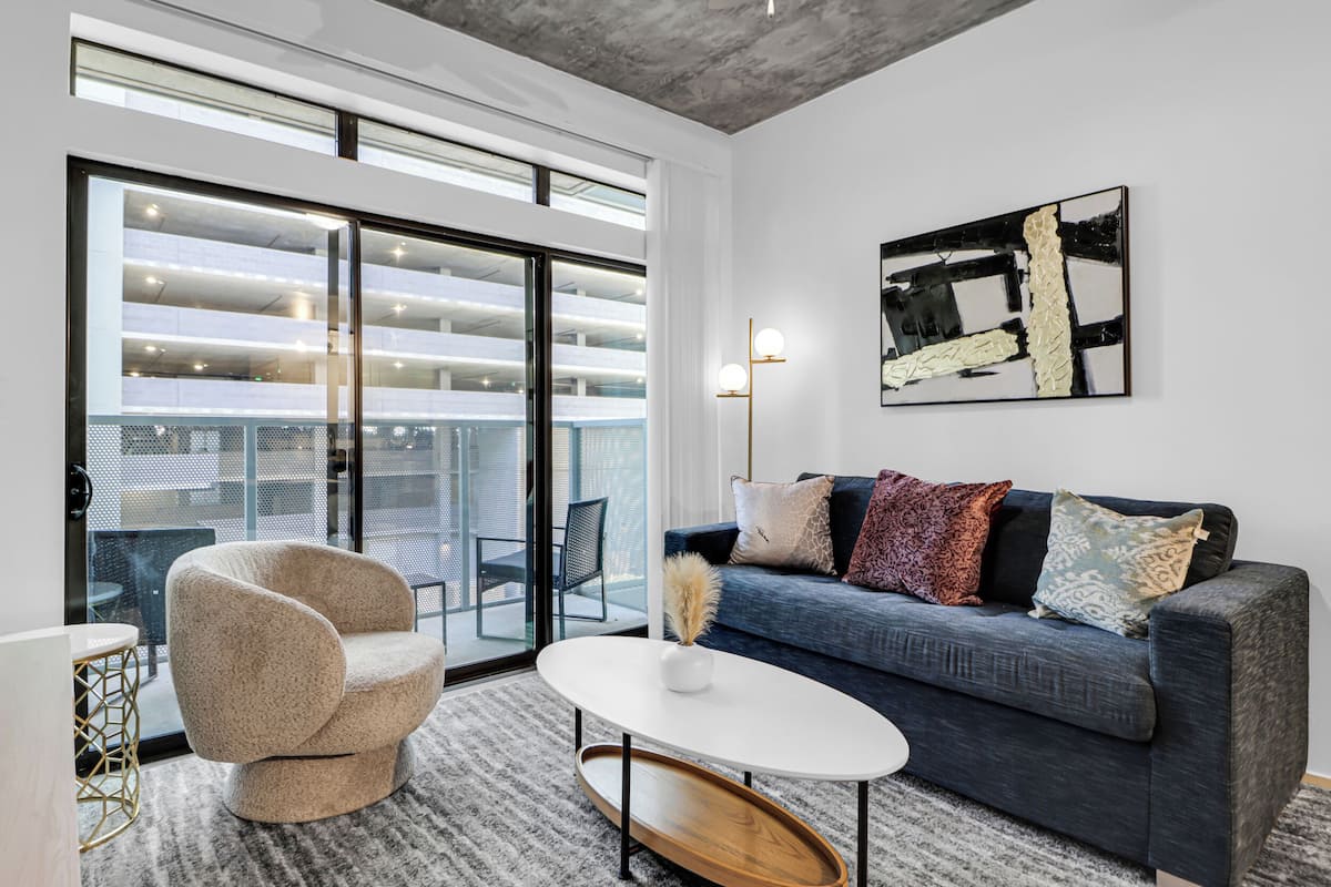 Ein modernes Wohnzimmer mit einem grauen Sofa, einem runden Couchtisch und einem beigen Sessel. Ein großes Fenster bietet Blick auf eine Parkgarage.