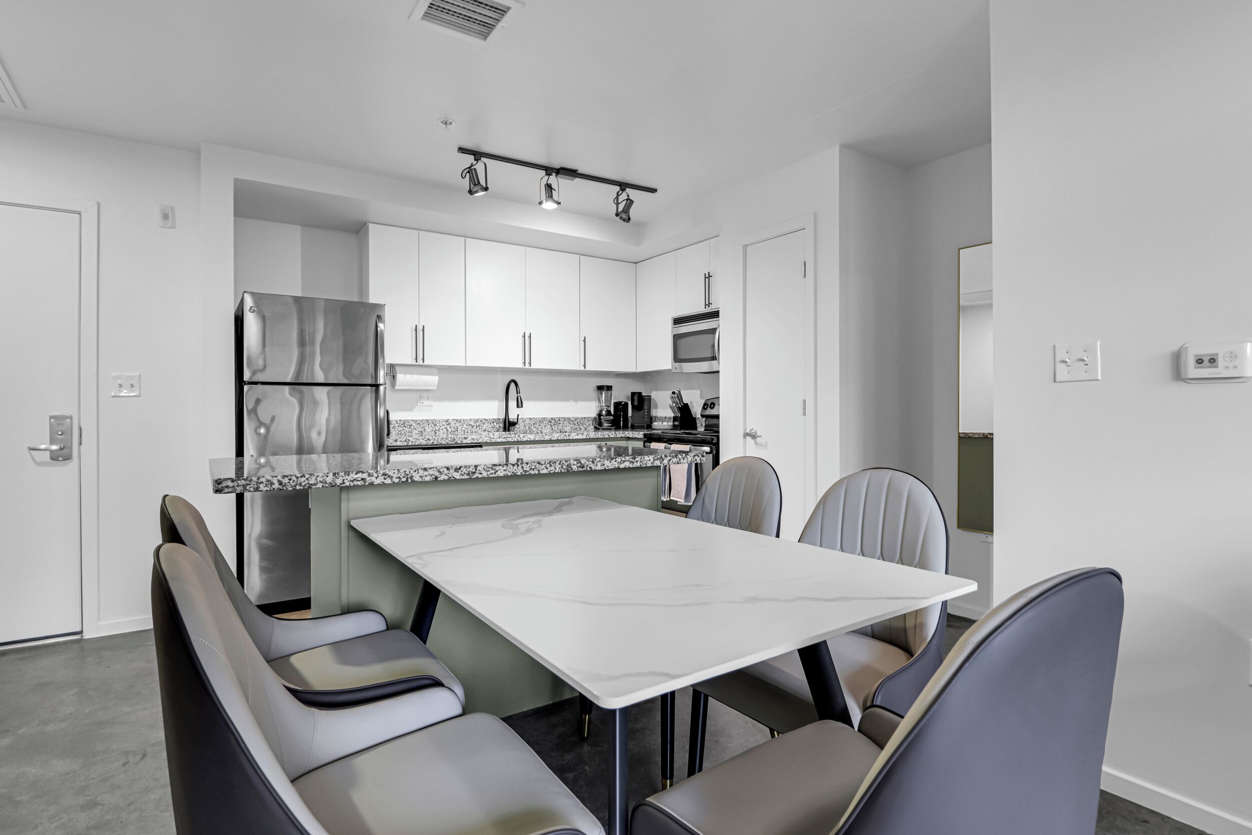 Eine moderne Küche mit weißen Schränken, einem Edelstahl-Kühlschrank, einer Mikrowelle und einem eingebauten Backofen. Es gibt einen Essbereich mit einem weißen Tisch und grauen Stühlen.