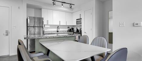 Eine moderne Küche mit weißen Schränken, einem Edelstahl-Kühlschrank, einer Mikrowelle und einem eingebauten Backofen. Es gibt einen Essbereich mit einem weißen Tisch und grauen Stühlen.