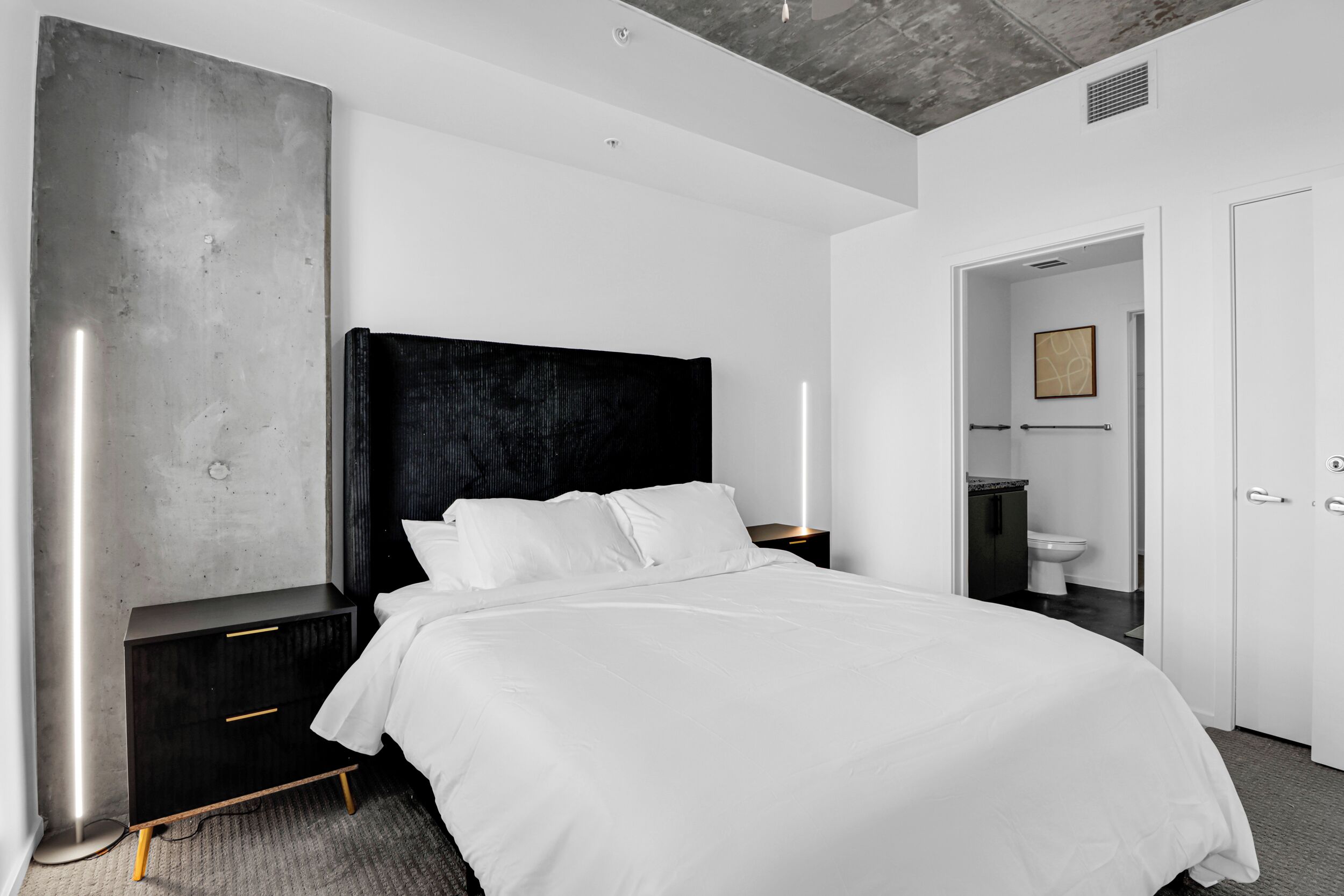 Une chambre moderne avec un grand lit, une tête de lit sombre, une table de chevet, une armoire et une salle de bain visible à travers une porte ouverte.