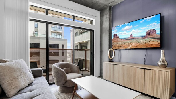 Un salon moderne comprenant un canapé, un fauteuil et une table basse, un téléviseur fixé au mur et une vue sur la ville grâce à de grandes fenêtres.