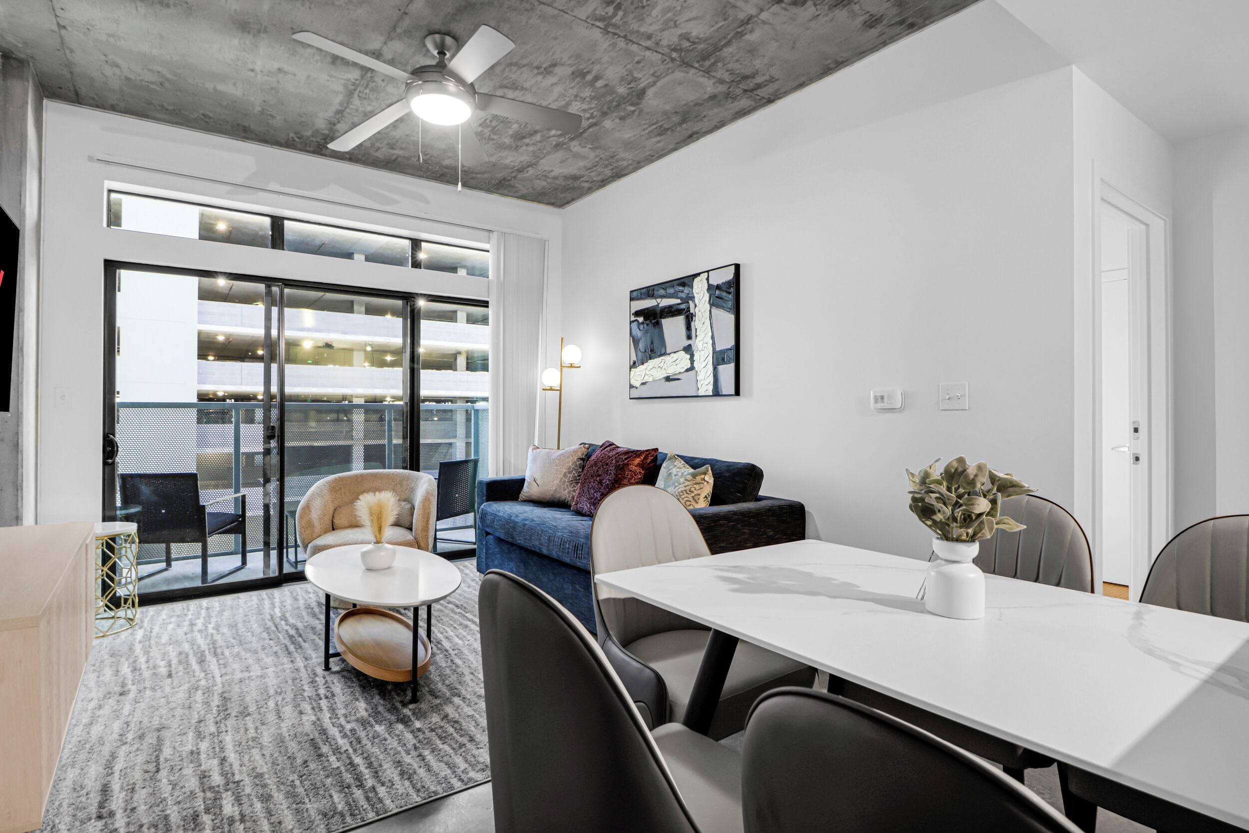 A modern living room with a white sofa, a round coffee table, and a dining table. There is a large window with a view of a parking garage.