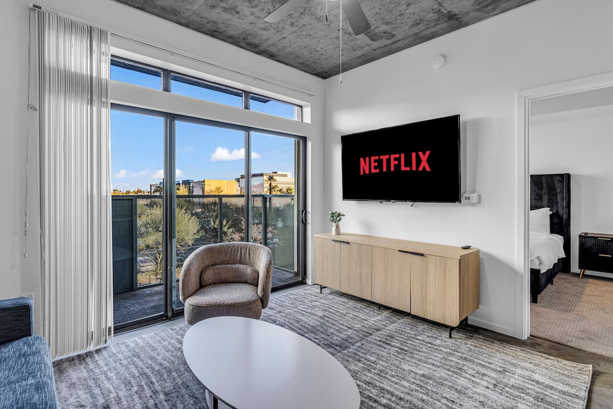 Ein modernes Wohnzimmer mit einem großen Fernseher, der "NETFLIX" anzeigt, einer bequemen Couch, einem kleinen Tisch und einer Schiebetür zu einem Balkon mit Blick auf die Stadt.