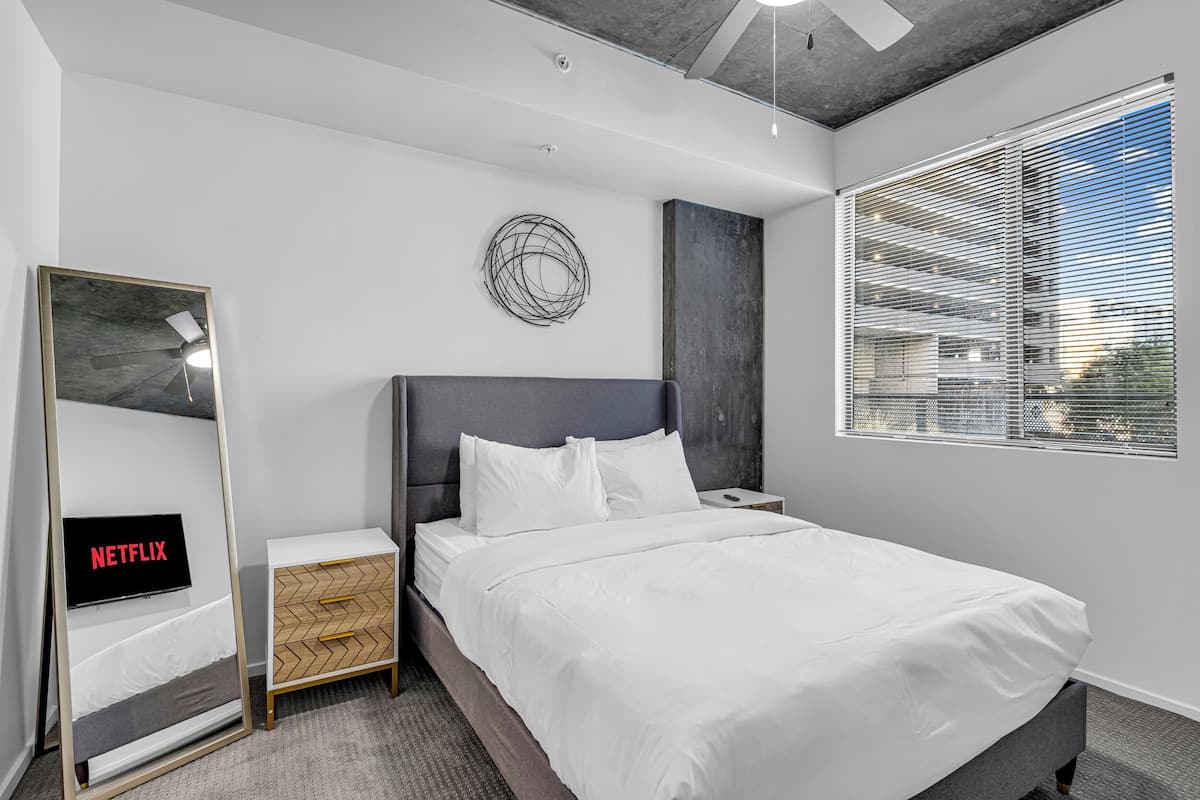 Ein modernes Hotelzimmer mit einem großen Bett, einem Nachttisch, einem an der Wand befestigten Fernseher und einem Fenster mit Jalousien.