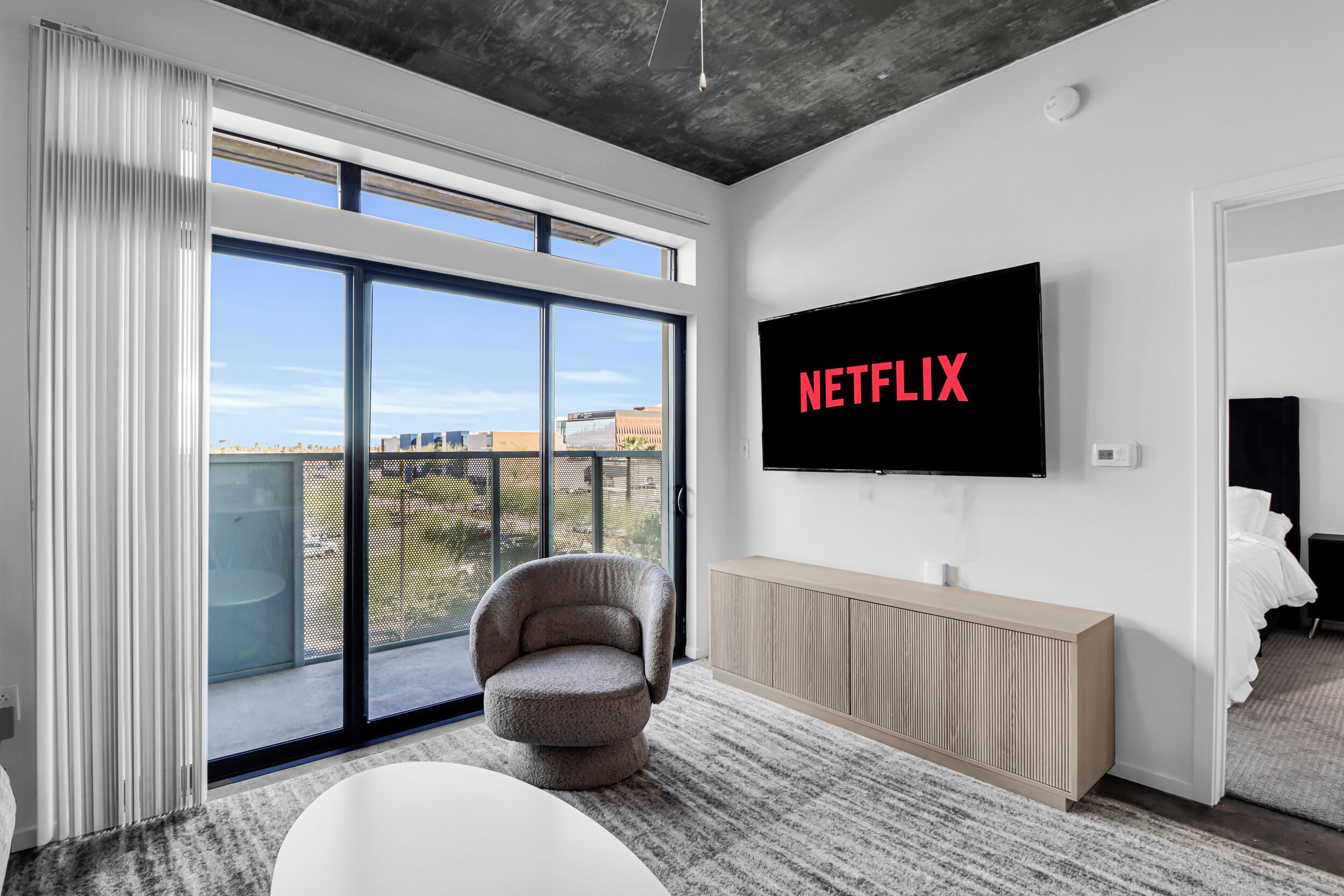 A modern living room with a large TV displaying "NETFLIX," a comfortable armchair, a white sofa, and a view through a sliding glass door.