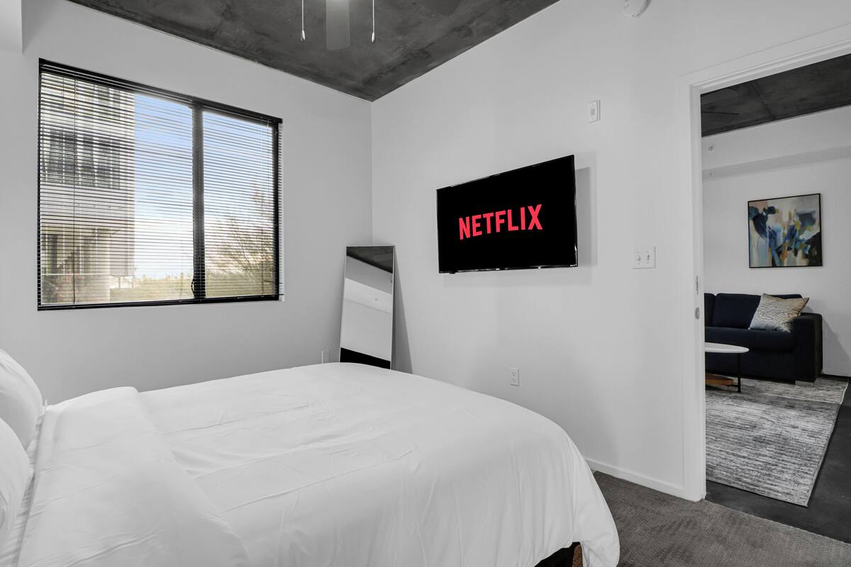 Ein modernes Schlafzimmer mit einem großen Bett, einem an der Wand montierten Fernseher und einem Fenster mit Jalousien.