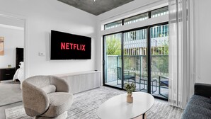 Un salon moderne avec une grande télévision affichant le logo « NETFLIX », un fauteuil gris, une table basse ronde ornée d’une plante, et une vue sur un balcon aménagé avec du mobilier d’extérieur.