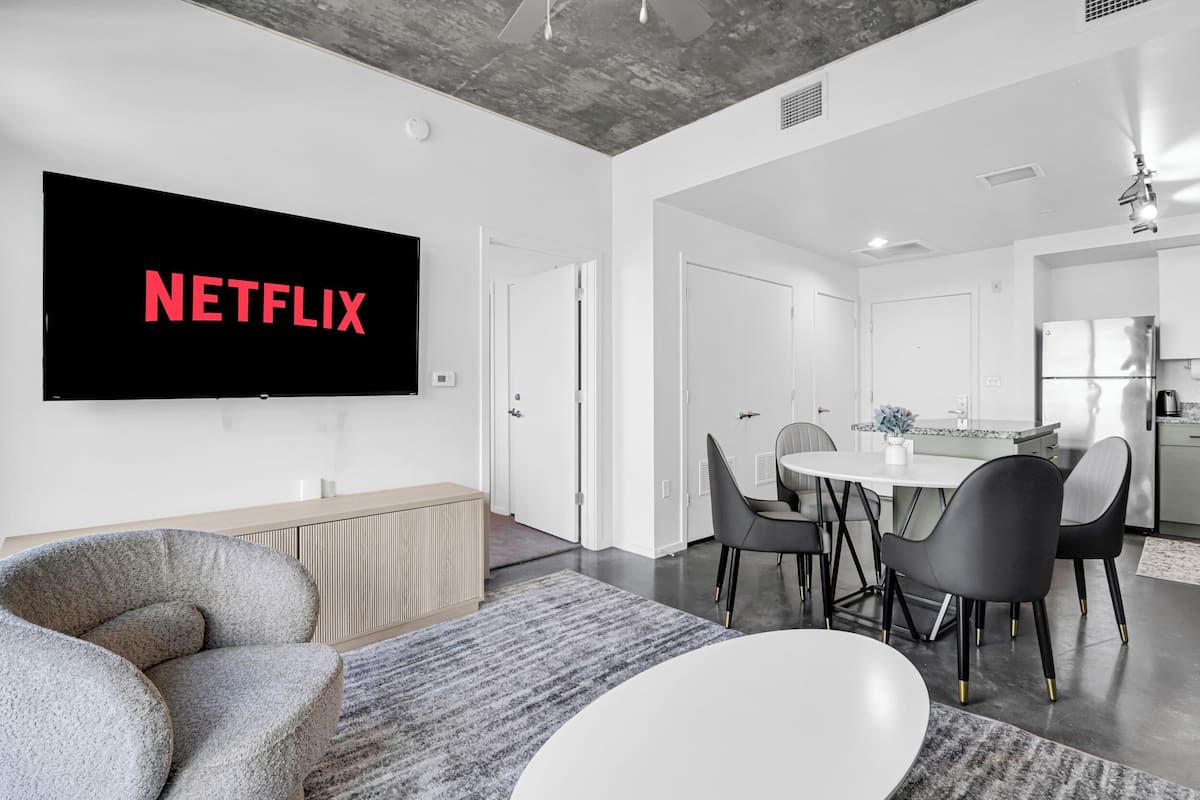 Ein modernes Wohnzimmer mit einem großen Flachbildfernseher, der "NETFLIX" anzeigt, einem Essbereich mit rundem Tisch und schwarzen Stühlen sowie einer Küche mit weißen Schränken und einem Kühlschrank.