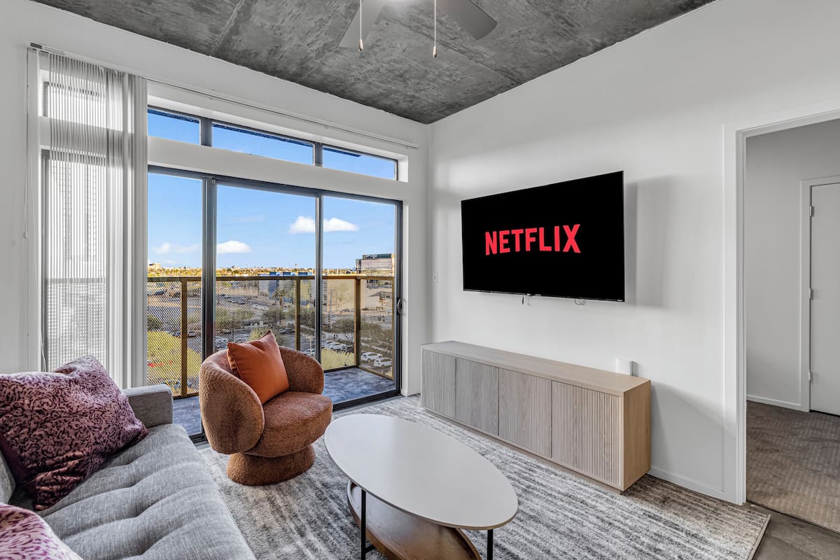 Ein modernes Wohnzimmer mit einem großen Fernseher, der "NETFLIX" anzeigt, einem grauen Sofa, einem braunen Sessel und einem großen Fenster mit Blick auf die Stadt.