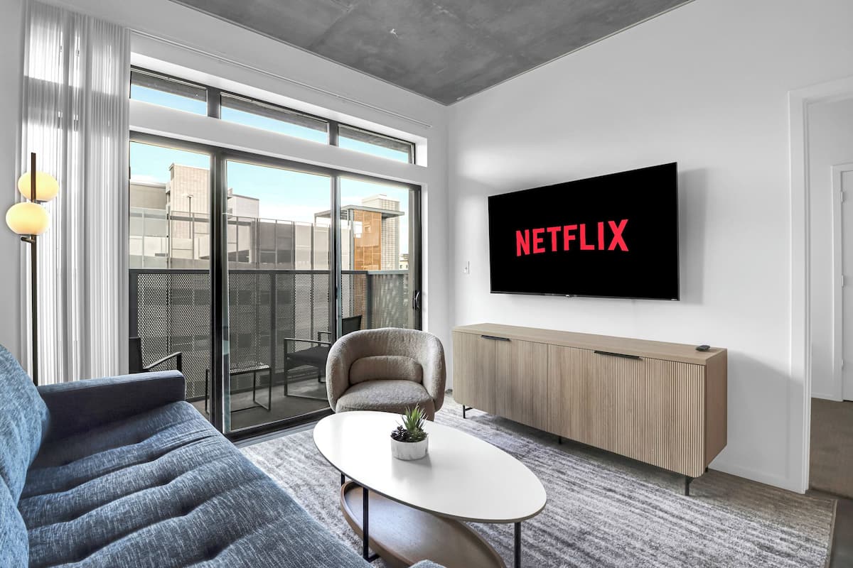 Ein modernes Wohnzimmer mit einem großen Fernseher, der "NETFLIX" anzeigt, einem Sofa, einem Couchtisch mit einer Pflanze und einer Glastür, die zu einem Außenbereich führt.