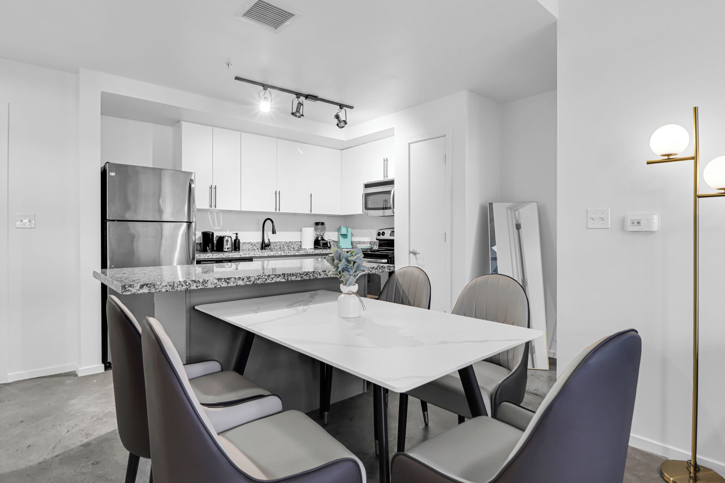 A modern kitchen with white cabinets, a stainless steel refrigerator, a built-in oven, and a granite countertop. There is a dining area with a white table and four chairs.