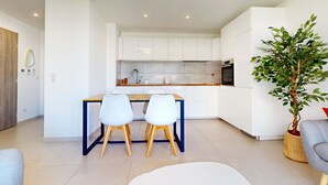 A modern kitchen with white cabinets, a wooden countertop, and a built-in oven. There is a dining area with a table and two chairs. A potted plant is in the corner.