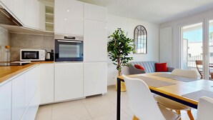 A modern kitchen with white cabinets, a microwave, and an oven. There is a dining area with a table, chairs, and a plant.