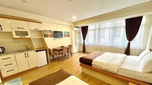 Ein kleines Hotelzimmer mit Bett, einer kompakten Küchenzeile, einem Essbereich und einem großen Fenster mit Vorhängen.