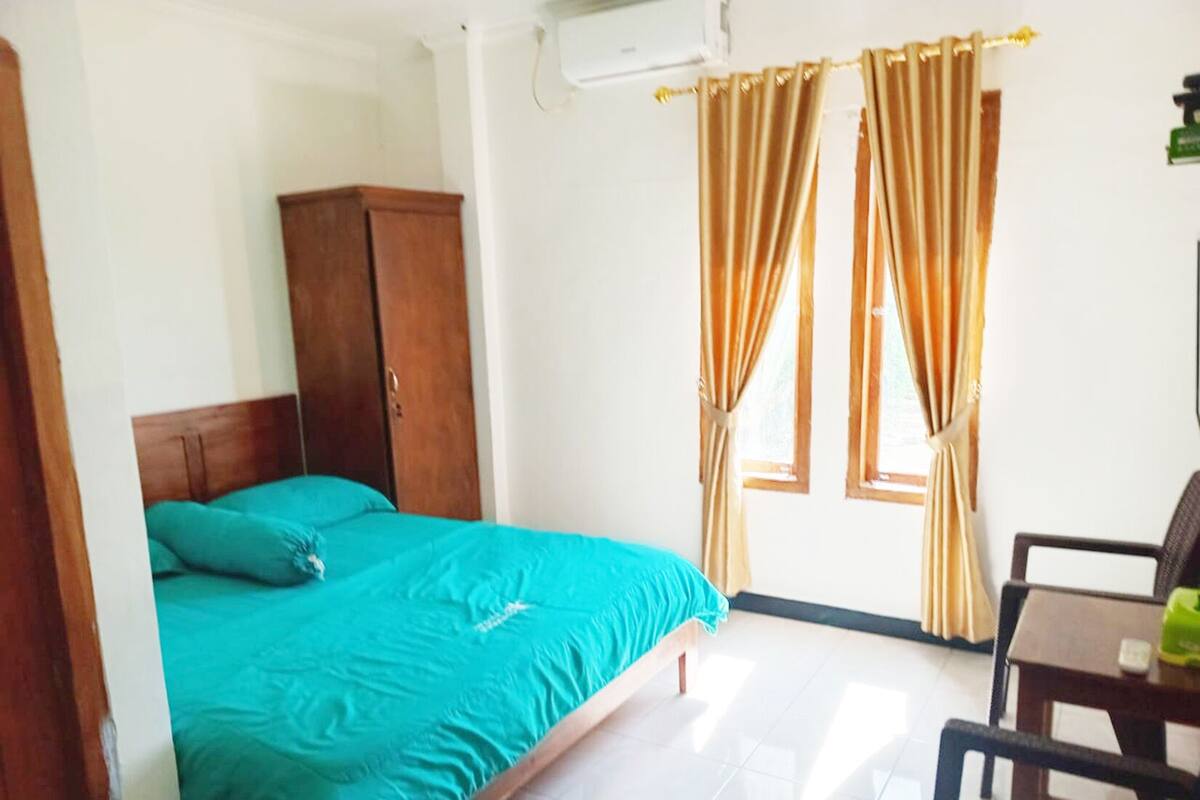 A bedroom with a bed, a wooden wardrobe, a window with curtains, and a small table with a chair.