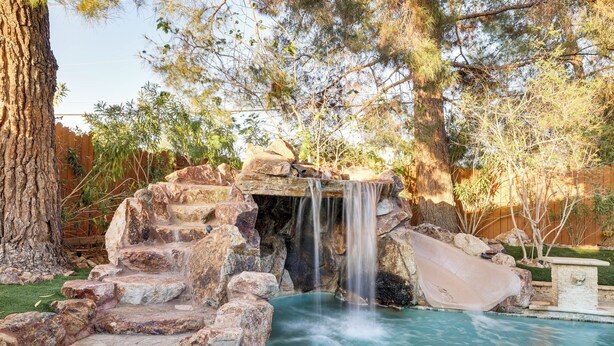 A pool with a waterfall feature and a slide, surrounded by rocks and trees. - 1-acre Las Vegas Getaway: Pool, Spa & Tennis Court (Las Vegas)