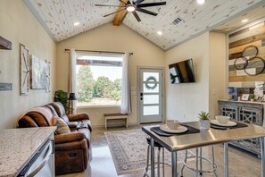 A spacious living area with a ceiling fan, a large window, a sofa, a dining table, and a kitchen area with a TV and decorative wall art.