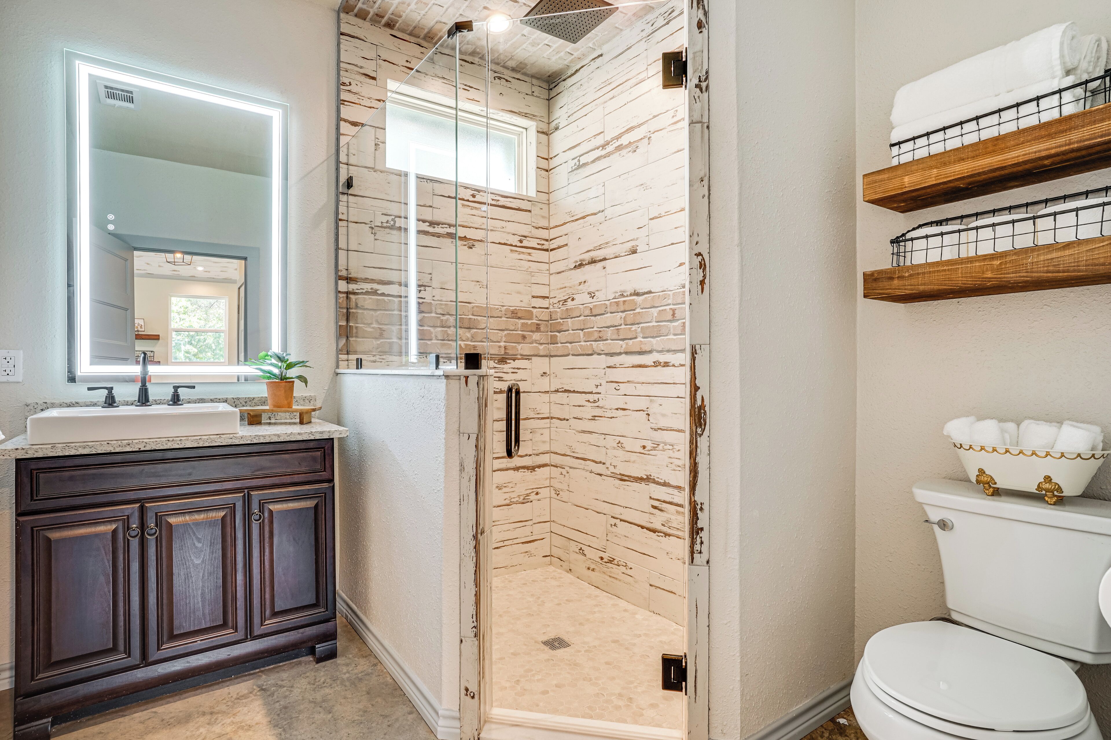 A bathroom with a glass-enclosed shower, a dark wooden vanity, a white toilet, and a mirror with a lighted frame.