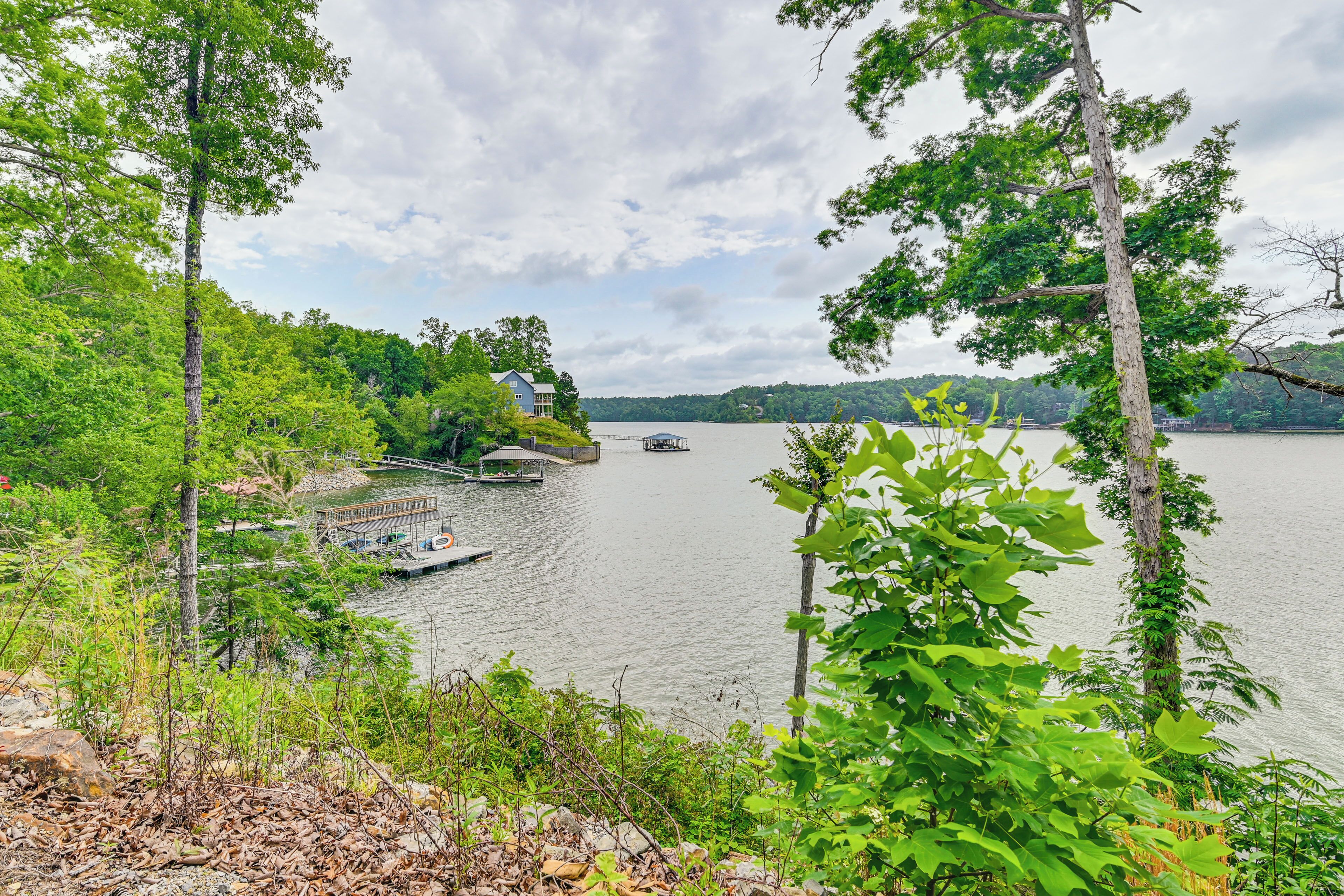 A lakeside property with a dock, surrounded by dense greenery and a house in the background.