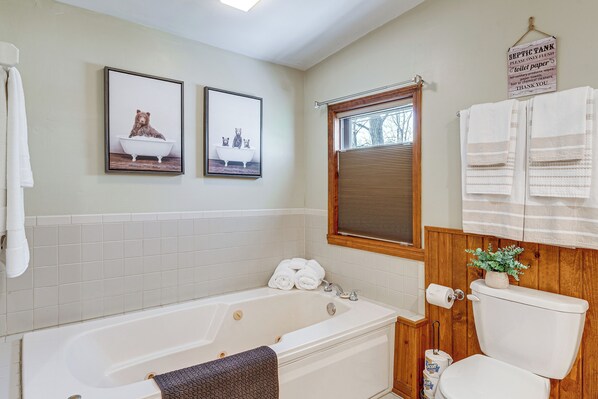 A bathroom with a bathtub, toilet, and a window with blinds. There are two framed pictures on the wall, a sign above the toilet, and a towel rack with towels.
