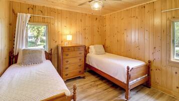 Een slaapkamer met houten lambrisering, twee bedden, een dressoir, een plafondventilator en een raam met gordijnen.