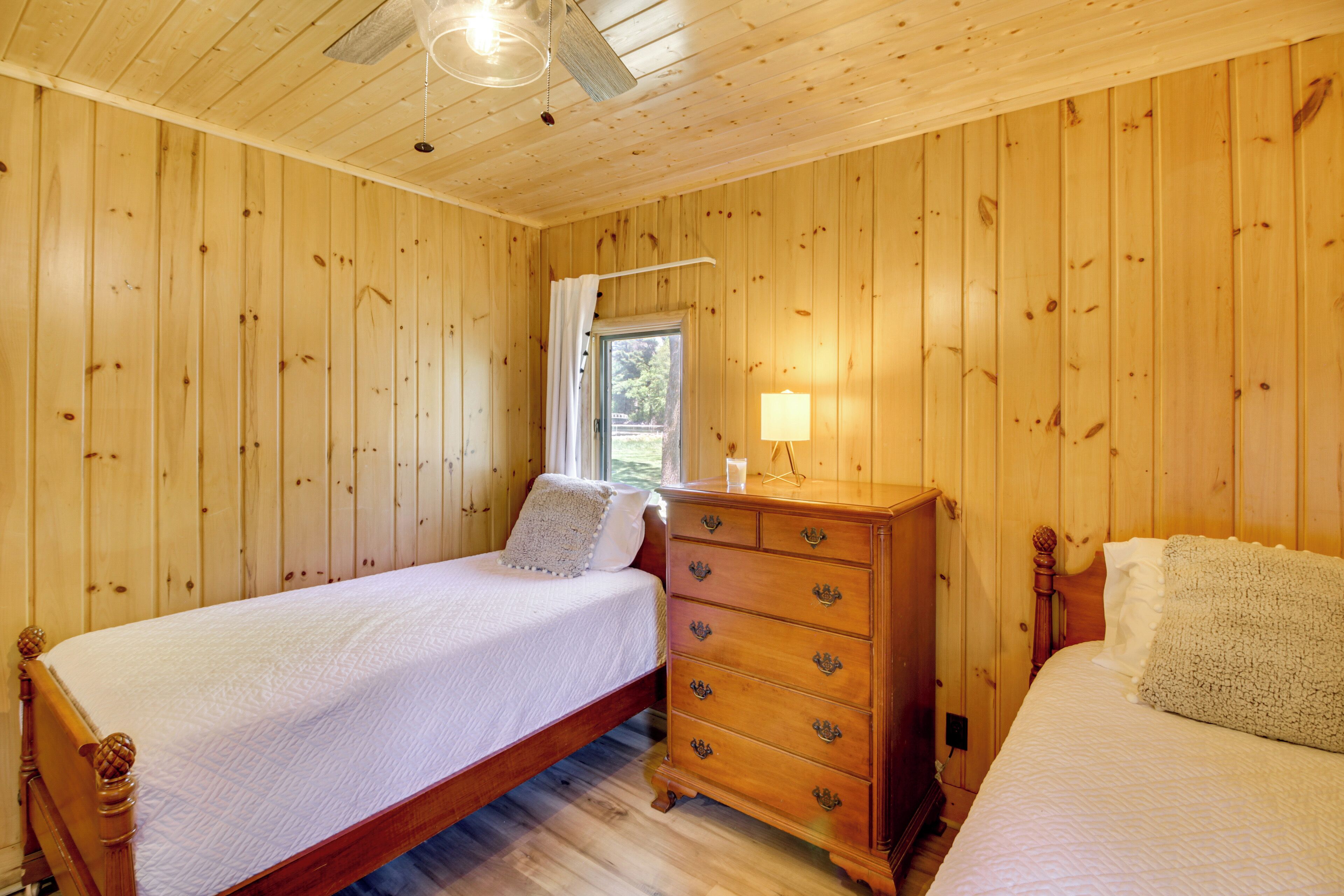 A wooden-paneled bedroom with two beds, a dresser, and a window with a curtain.